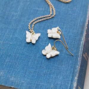 May include: A delicate gold chain necklace and matching earrings feature white butterfly charms. The charms have a sparkly, iridescent finish.