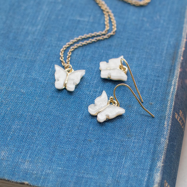 May include: A delicate gold chain necklace and matching earrings feature white butterfly charms. The charms have a sparkly, iridescent finish.