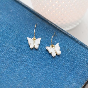May include: A pair of gold butterfly earrings with white butterfly charms. The earrings are on a blue surface.