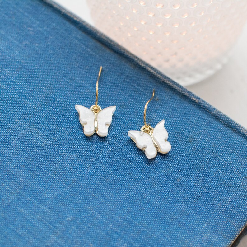May include: A pair of gold butterfly earrings with white butterfly charms. The earrings are on a blue surface.