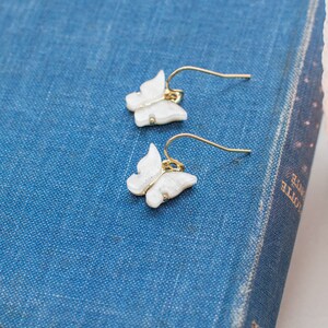 May include: A pair of gold butterfly earrings with white glitter accents. The earrings are on a blue surface.