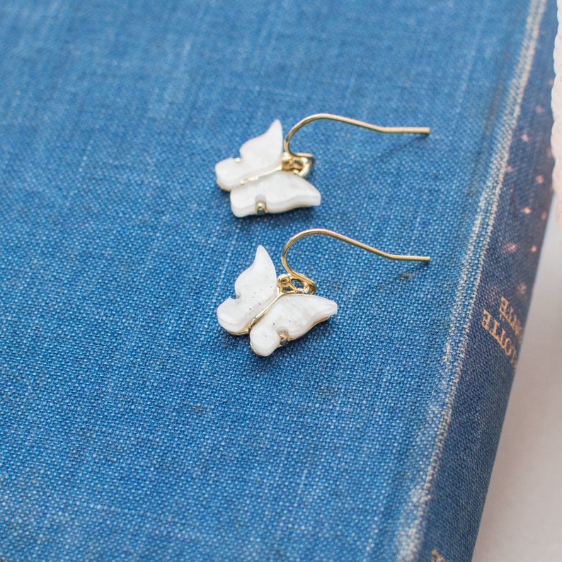 May include: A pair of gold butterfly earrings with white glitter accents. The earrings are on a blue surface.