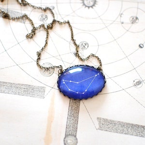 May include: A silver chain necklace with a blue glass pendant featuring a white constellation design. The pendant is oval shaped and has a silver rim.