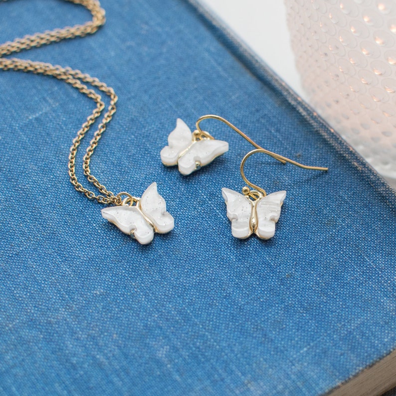 May include: A gold chain necklace and a pair of earrings with white butterfly charms. The butterfly charms have a sparkly, iridescent finish.