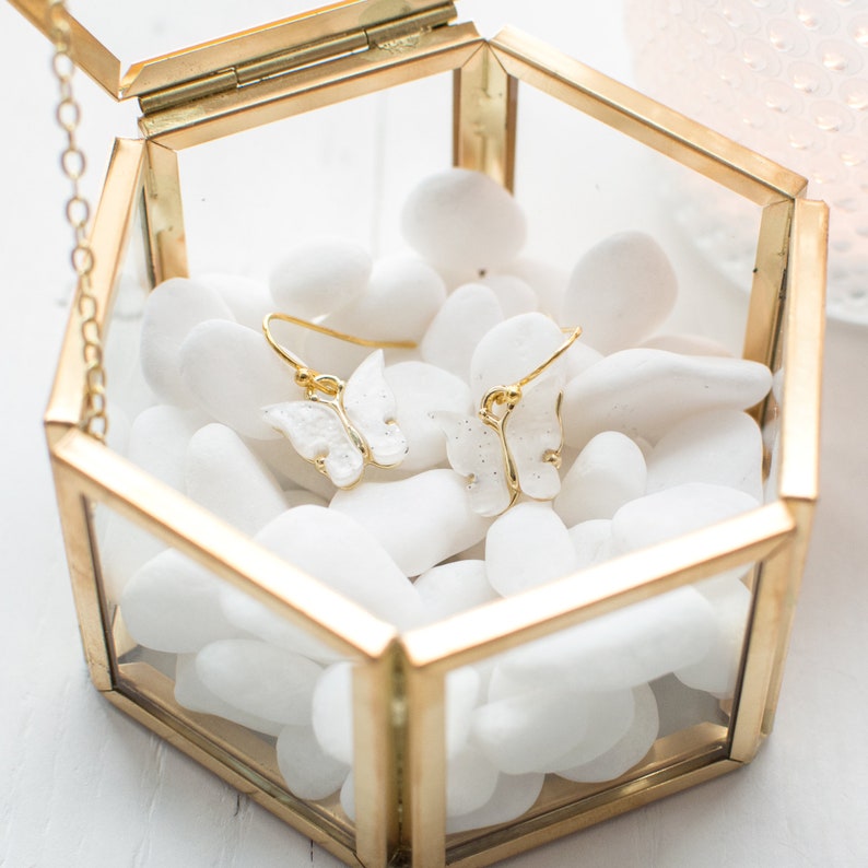 May include: A pair of white butterfly earrings with gold accents are displayed in a glass hexagonal box filled with white stones.
