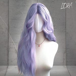 May include: A long, wavy lavender wig displayed on a mannequin head. The wig features a natural-looking hairline and soft, flowing curls. A delicate necklace with a pink pendant adorns the mannequin's neck. The word "LIORA" is written in cursive in the top right corner.