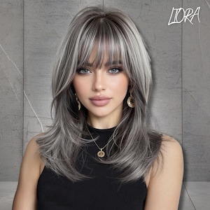 May include: A shoulder-length wig featuring layered, ash-gray hair with darker roots and straight-cut bangs. The hair has a multi-tonal appearance with lighter silver highlights throughout. The word LIORA is printed in white in the top right corner.