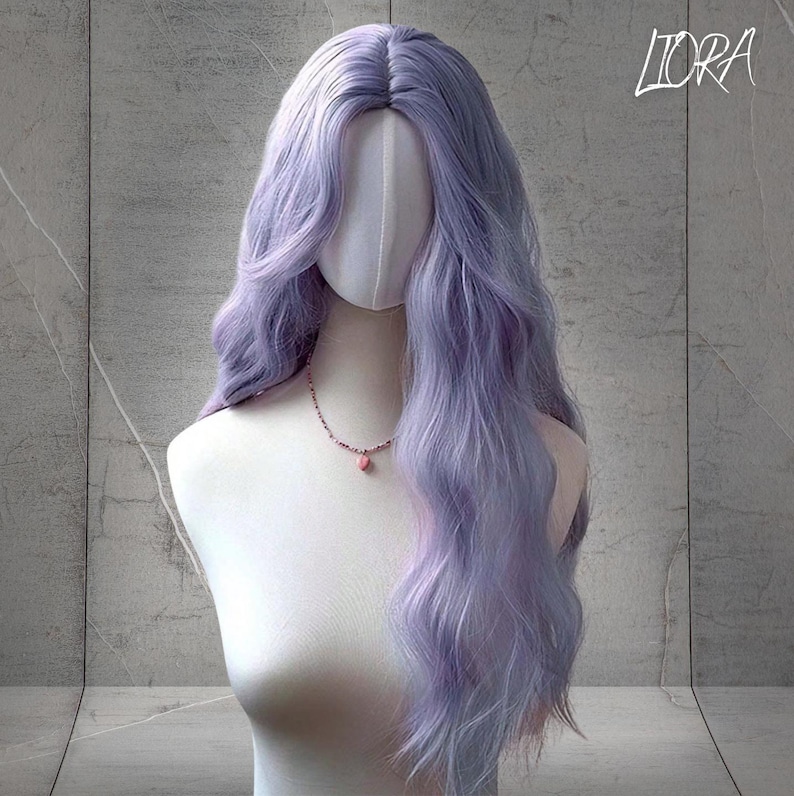 May include: A long, wavy lavender wig styled on a mannequin head. The wig features a natural-looking part and soft, flowing layers. A delicate pink necklace with a pendant adorns the mannequin's neck. The word "LIORA" is written in cursive in the top right corner.