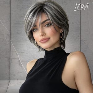 May include: A short, layered wig featuring a blend of silver, gray, and dark brown hair with side-swept bangs. The hair is styled in a textured, chin-length cut. The word LIORA is printed in white in the upper right corner of the image.