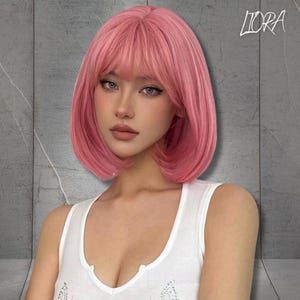 Può includere: Una parrucca bob rosa con frangia. La parrucca è di una tonalità rosa vibrante e ha una consistenza liscia e liscia. I capelli sono acconciati in un bob fino al mento con una frangia piena. La parrucca è indossata da una persona che indossa una canotta bianca. La parola "LIORA" è scritta in alto a destra.
