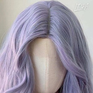 May include: A lavender-colored wig with long, wavy hair. The wig features a natural-looking part and a blend of purple and gray tones. The text "LIORA" is visible in the upper right corner.