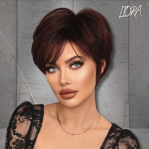 May include: A short, layered auburn wig with side-swept bangs. The wig is styled with a slight lift at the crown and is worn by a person with fair skin, blue eyes, and full lips. A gold necklace is visible. The word "LIORA" is in the upper right corner.
