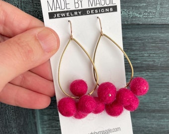 Może przedstawiać: Złote kolczyki koła z wiszącymi różowymi kulkami z filcu. Kolczyki znajdują się na białej kartce z tekstem "Made by Maggie Jewelry Designs" i "www.madebymaggie.com".