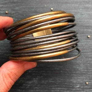 May include: A gold and silver leather bracelet with multiple bands. The bracelet is made of thin leather strips that are wrapped around each other.