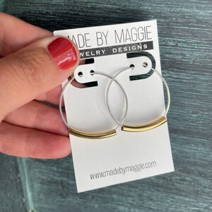 May include: Silver hoop earrings with a gold bar accent. The earrings are displayed on a white card with the text "MADE BY MAGGIE JEWELRY DESIGNS" and the website address www.madebymaggie.com.
