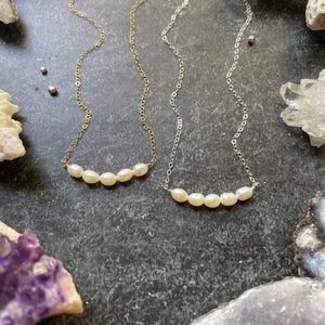 Puede incluir: Dos delicados collares con una fila de cinco perlas blancas cada uno. Los collares est&aacute;n sobre un fondo gris oscuro con varias piedras y conchas.