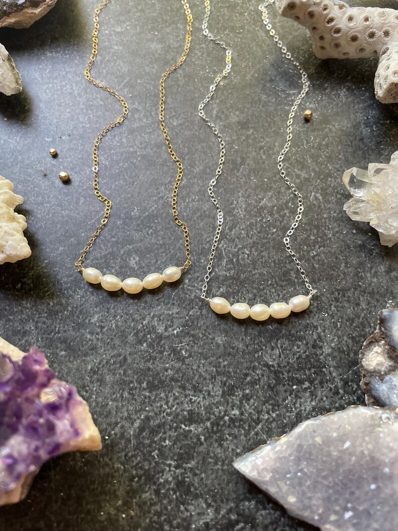 Puede incluir: Dos delicados collares con una fila de cinco perlas blancas cada uno. Los collares est&aacute;n sobre un fondo gris oscuro con varias piedras y conchas.