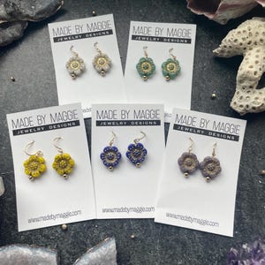 May include: Five pairs of floral earrings in various colors, including yellow, blue, green, and gray, displayed on white cards. Each card features the text "MADE BY MAGGIE JEWELRY DESIGNS" and the website address. The earrings have gold-colored hooks.