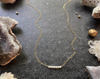 May include: A delicate gold chain necklace with a row of small white pearls. The necklace is displayed on a dark surface with various natural elements.