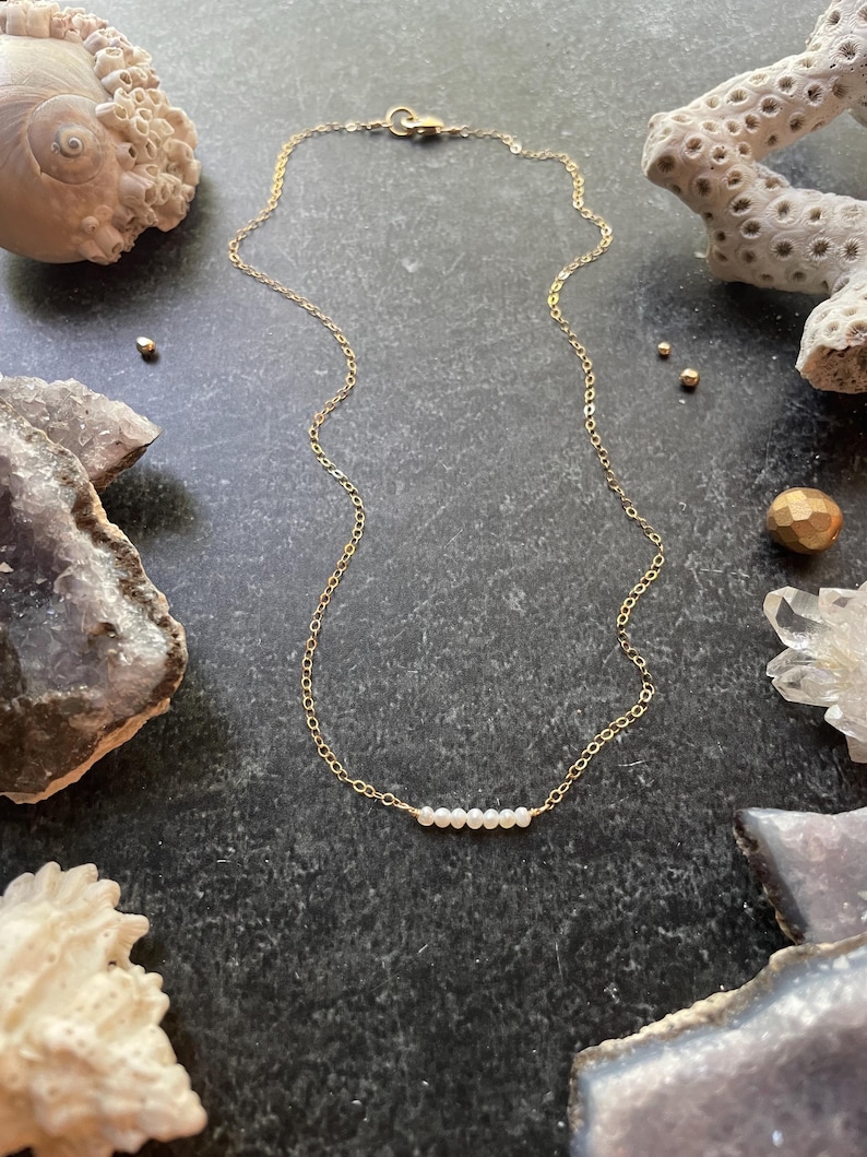 May include: A delicate gold chain necklace with a row of small white pearls. The necklace is displayed on a dark surface with various natural elements.