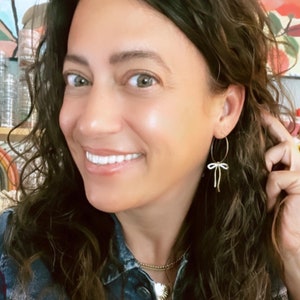 May include: A woman wearing a denim shirt with a gold chain necklace and hoop earrings with a white bow charm.