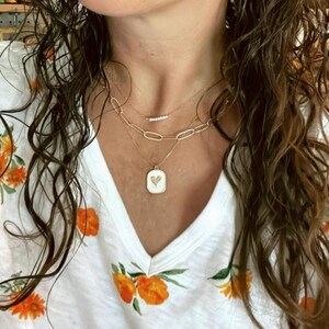 May include: A woman wearing a white shirt with orange floral print and a gold chain necklace with a small heart pendant. The necklace is layered with two other gold chains.