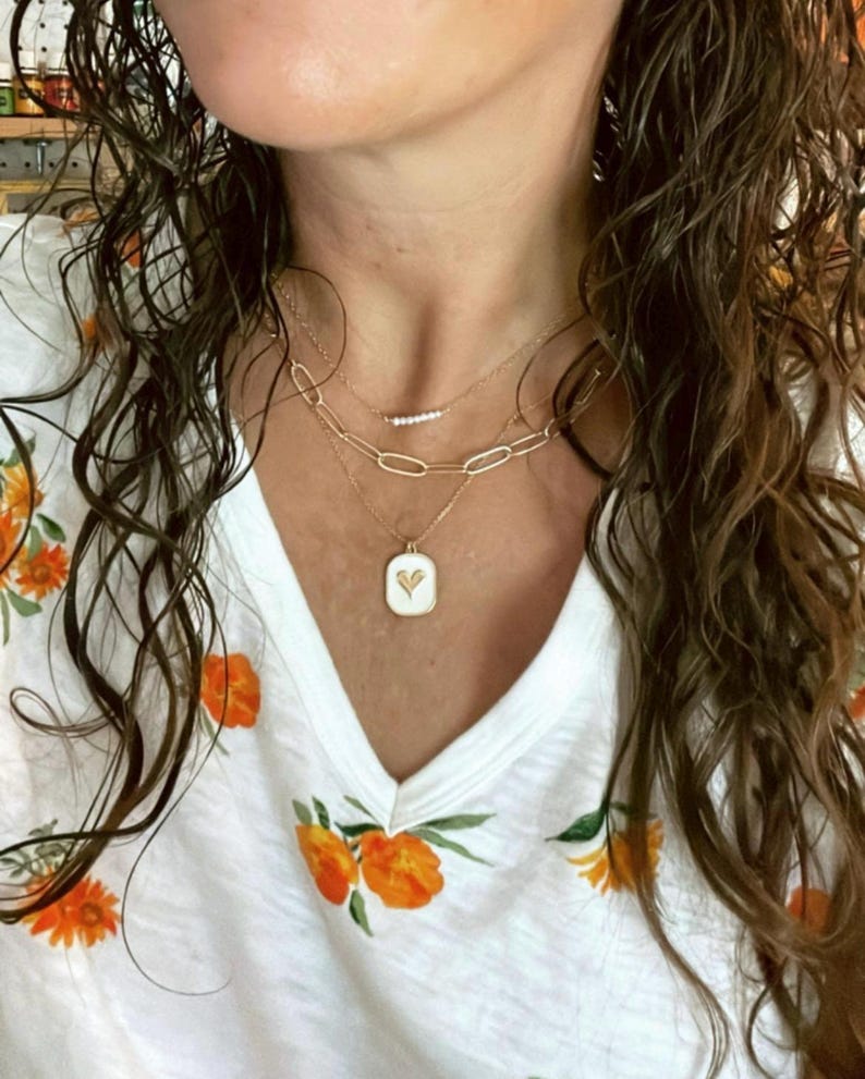 May include: A woman wearing a white shirt with orange floral print and a gold chain necklace with a small heart pendant. The necklace is layered with two other gold chains.