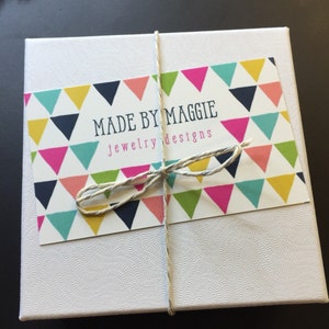 May include: A white box with a colorful label that says "Made by Maggie jewelry designs". The label has a pattern of colorful triangles.