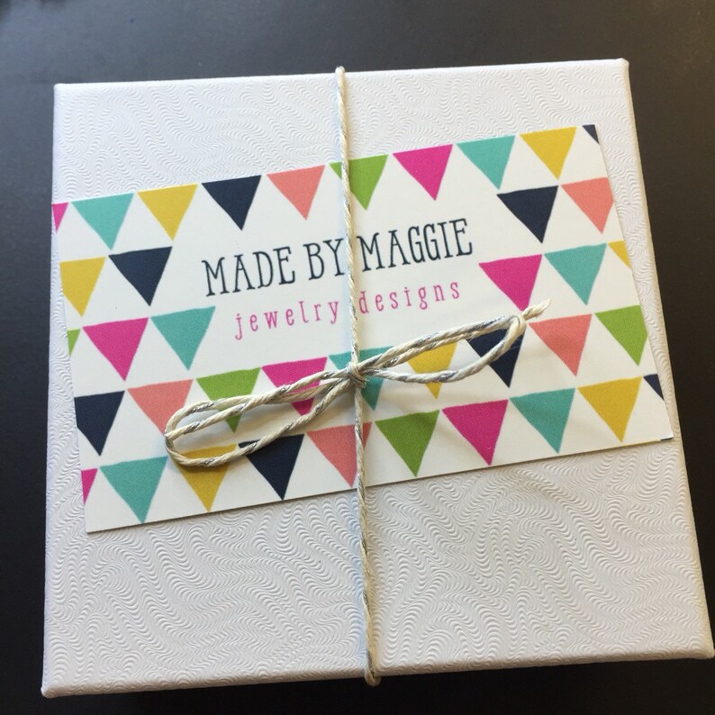 May include: A white box with a colorful label that says "Made by Maggie jewelry designs". The label has a pattern of colorful triangles.