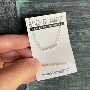 May include: A delicate gold chain necklace with a row of small white pearls. The necklace is displayed on a white card with the text "Made by Maggie Jewelry Designs" and the website address "www.madebymaggie.com".