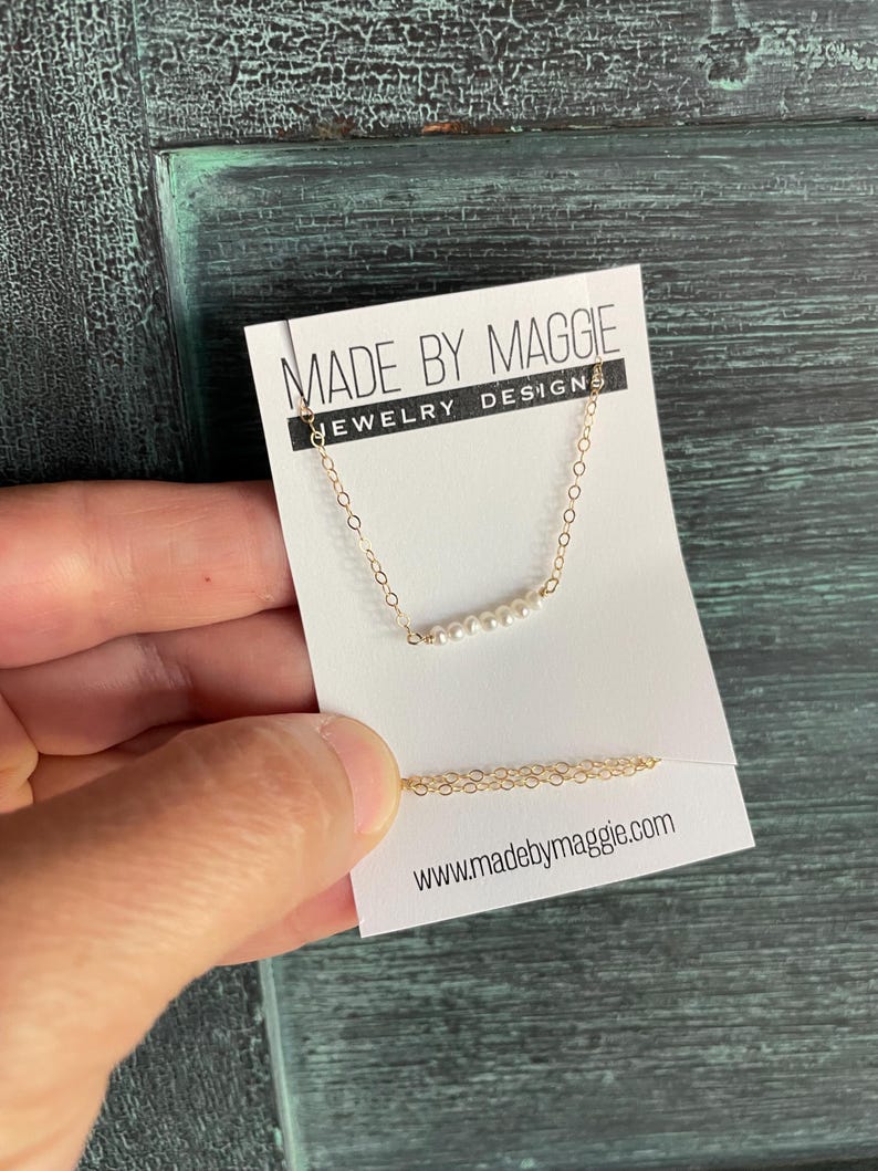 May include: A delicate gold chain necklace with a row of small white pearls. The necklace is displayed on a white card with the text "Made by Maggie Jewelry Designs" and the website address "www.madebymaggie.com".