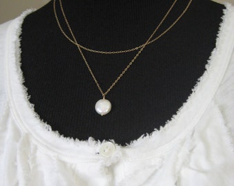 May include: A delicate gold-tone necklace with two strands. The lower strand features a large, round, white pearl pendant. The necklace is displayed on a black mannequin, against a white, textured top.