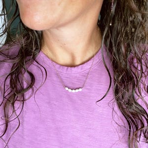 Puede incluir: Una mujer con una camisa morada con un delicado collar de cadena dorada con una fila de peque&ntilde;as perlas blancas.