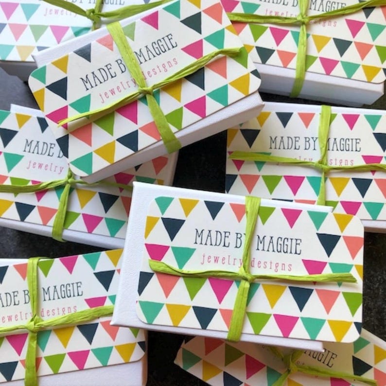Puede incluir: Varias cajas blancas con patrones geom&eacute;tricos coloridos y una cinta verde. Cada caja tiene una etiqueta que dice "Made by Maggie Jewelry Designs".
