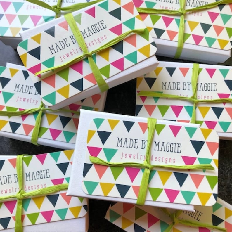 K&ouml;nnte beinhalten: Mehrere wei&szlig;e Schmuckschatullen sind angeordnet, jede mit einem farbenfrohen Dreiecksmuster auf dem Deckel. Die Schachteln sind mit einem hellgr&uuml;nen Band gebunden. Der Text "MADE BY MAGGIE jewelry designs" ist auf dem Deckel aufgedruckt.