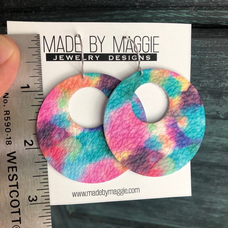 May include: A pair of tie-dye leather hoop earrings with a silver hoop. The earrings are approximately 2 inches in diameter and feature a colorful tie-dye pattern in shades of pink, blue, yellow, and purple. The earrings are made by Made By Maggie Jewelry Designs.