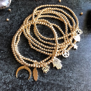 May include: A set of seven gold-toned beaded bracelets with various charms, including a crescent moon, a feather, a lotus flower, an evil eye, a hamsa hand, a peace sign, a star, and a heart.