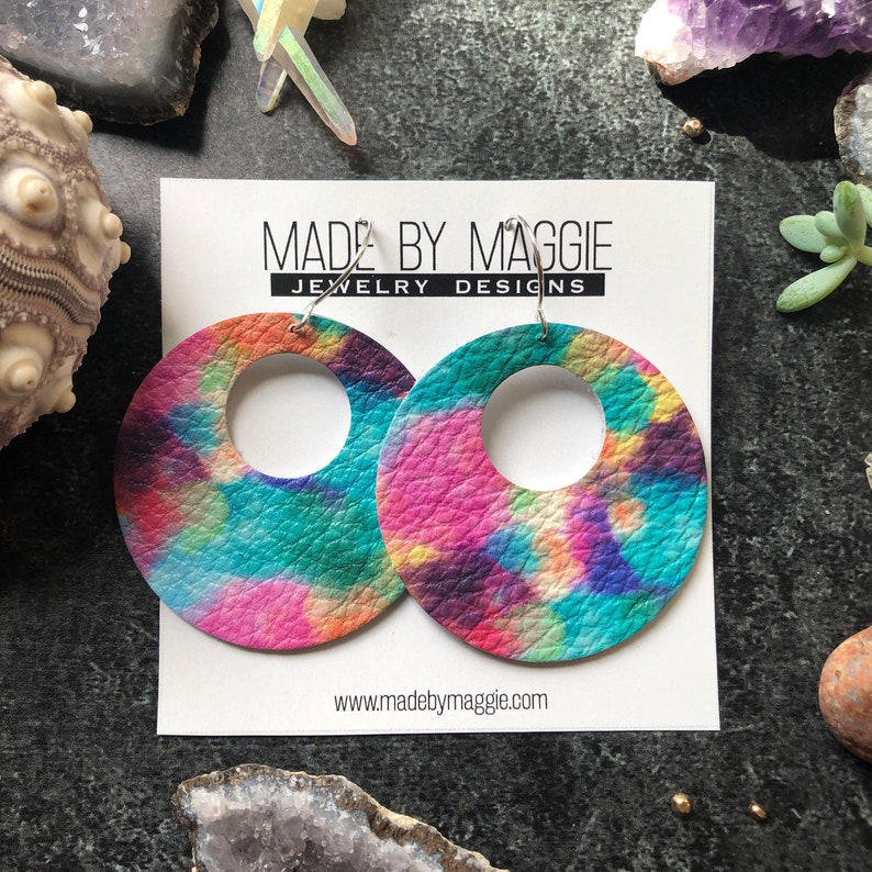 May include: A pair of large, round, tie-dye earrings with a hole in the center. The earrings are made of leather and have a pink, blue, green, and yellow color scheme. The earrings are on a white card with the text "MADE BY MAGGIE JEWELRY DESIGNS" and "www.madebymaggie.com".