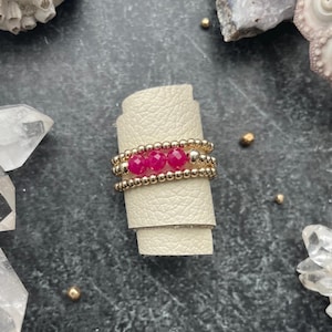 May include: A set of three gold beaded stacking rings with pink gemstone accents. The rings are stacked on a white leather surface.