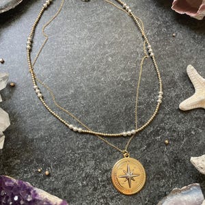 K&ouml;nnte beinhalten: Eine goldfarbene Halskette mit einem Kompassanh&auml;nger, der ein Sterndesign aufweist. Die Halskette hat zwei Str&auml;nge, einer mit kleinen Perlen. Der Anh&auml;nger ist rund mit einer detaillierten Windrose. Der Hintergrund umfasst Kristalle und Muscheln.