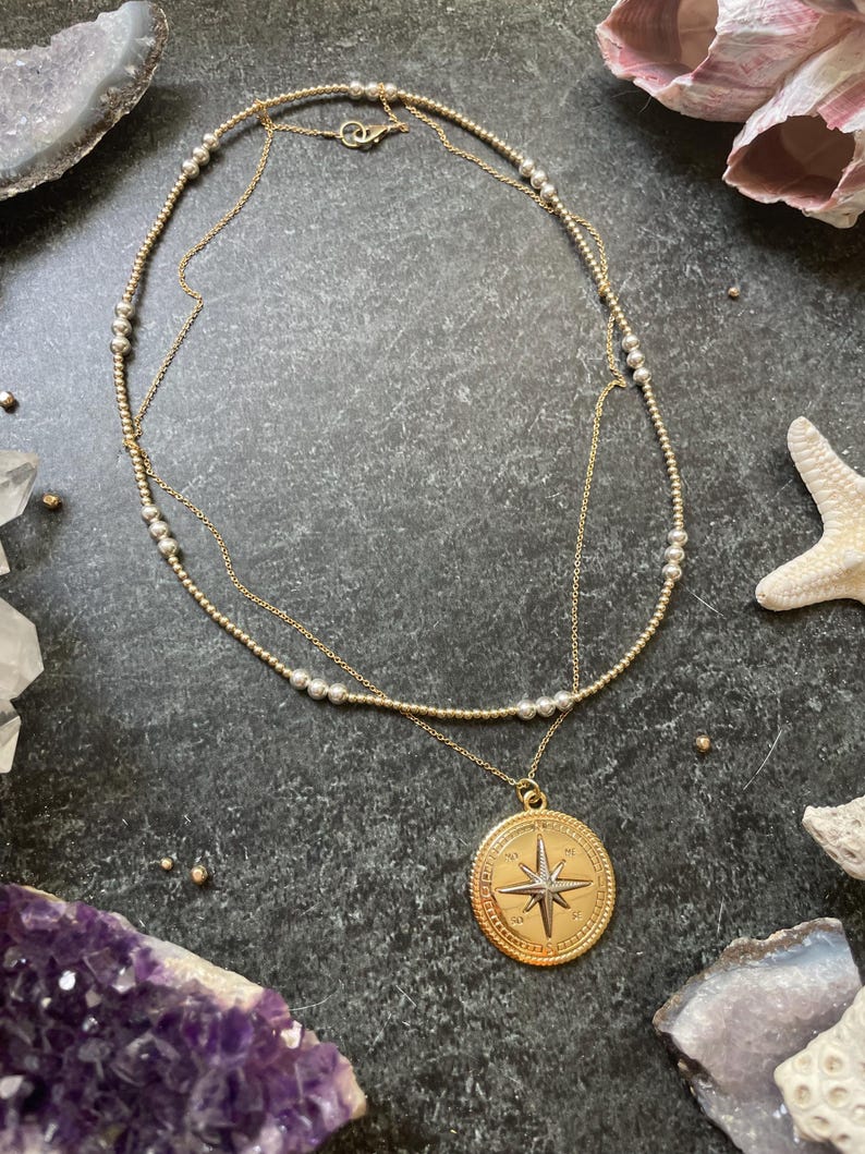 K&ouml;nnte beinhalten: Eine goldfarbene Halskette mit einem Kompassanh&auml;nger, der ein Sterndesign aufweist. Die Halskette hat zwei Str&auml;nge, einer mit kleinen Perlen. Der Anh&auml;nger ist rund mit einer detaillierten Windrose. Der Hintergrund umfasst Kristalle und Muscheln.