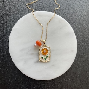 May include: Gold-tone necklace featuring a rectangular pendant with a sunflower design. The pendant has a gold frame and a pearl in the center. The necklace also includes a small orange bead and a pearl charm.