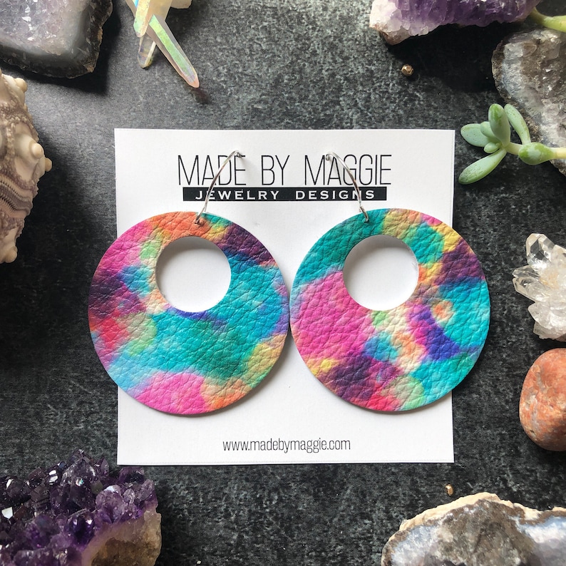 May include: A pair of colorful tie-dye leather earrings with a large circle design. The earrings are made by Made By Maggie Jewelry Designs.