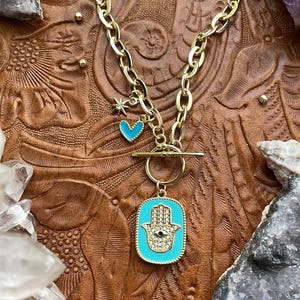 May include: A gold chain necklace with a turquoise blue hamsa hand charm with a white evil eye and a small blue heart charm. The necklace is on a brown leather background.