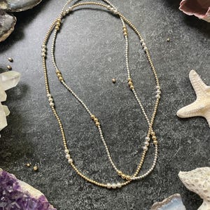 K&ouml;nnte beinhalten: Zwei zarte Halsketten mit abwechselnd goldenen und silbernen Perlen. Die Halsketten sind geschichtet und erzeugen einen stilvollen und eleganten Look. Der Schmuck wird auf einer dunklen, strukturierten Oberfl&auml;che mit dekorativen Elementen im Hintergrund pr&auml;sentiert.