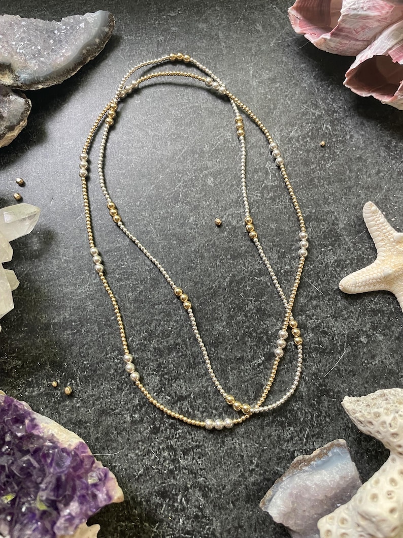 K&ouml;nnte beinhalten: Zwei zarte Halsketten mit abwechselnd goldenen und silbernen Perlen. Die Halsketten sind geschichtet und erzeugen einen stilvollen und eleganten Look. Der Schmuck wird auf einer dunklen, strukturierten Oberfl&auml;che mit dekorativen Elementen im Hintergrund pr&auml;sentiert.