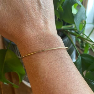 May include: A thin, smooth, gold-colored metal bangle bracelet worn around the wrist. The bracelet features a simple, circular design with a polished finish, maintaining a consistent, slender width throughout its entire circumference.