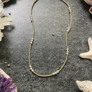 K&ouml;nnte beinhalten: Eine zarte Halskette mit abwechselnd gold- und silberfarbenen Perlen. Die Halskette besteht aus kleinen, runden Perlen, die zu einem einfachen, aber eleganten Design aufgereiht sind. Die Halskette wird auf einer dunklen, strukturierten Oberfl&auml;che mit dekorativen Elementen im Hintergrund pr&auml;sentiert.