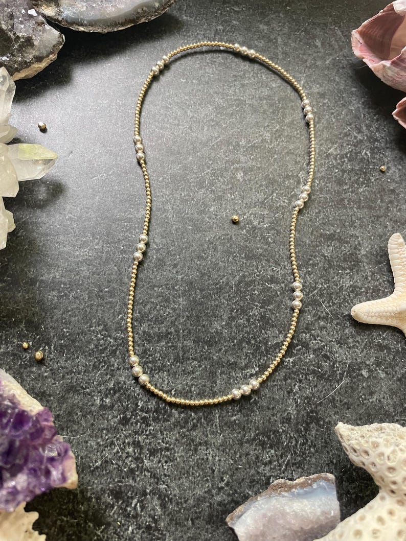 K&ouml;nnte beinhalten: Eine zarte Halskette mit abwechselnd gold- und silberfarbenen Perlen. Die Halskette besteht aus kleinen, runden Perlen, die zu einem einfachen, aber eleganten Design aufgereiht sind. Die Halskette wird auf einer dunklen, strukturierten Oberfl&auml;che mit dekorativen Elementen im Hintergrund pr&auml;sentiert.