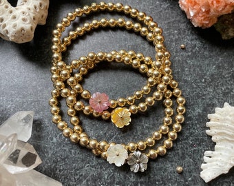 Puede incluir: Cuatro pulseras de cuentas doradas con dijes de flores. Los dijes están hechos de madreperla y vienen en rosa, amarillo, blanco y gris.